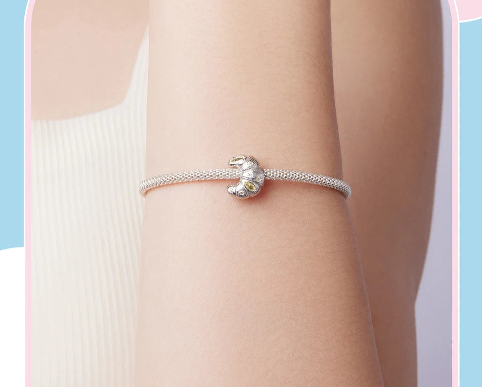 Silver bracelet with a croissant charm worn on a wrist against a light blue background